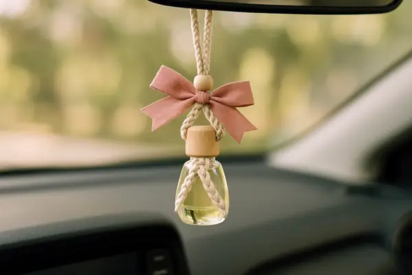 Aromatizador natural artesanal pendurado no retrovisor de um carro, com frasco de vidro, tampa de madeira, macramê e laço rosa.