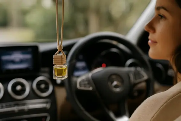 Difusor de óleo essencial pendurado no retrovisor de um carro com interior elegante, ao fundo uma mulher sorridente ao volante.